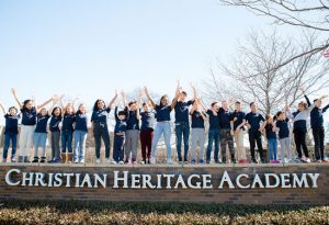 Over 20 K-12 students standing side by side with arms raised on a long brick wall sign that reads Christian Heritage Academy